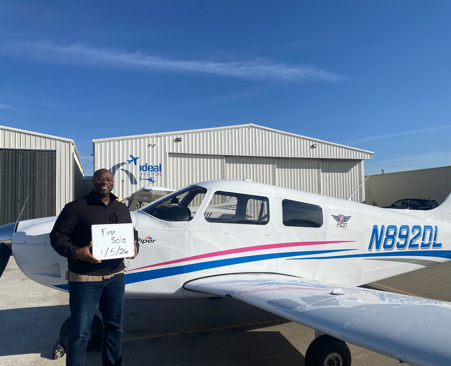 Ideal Aviation student holding a First Solo sign beside a Piper airplane outside the hangar